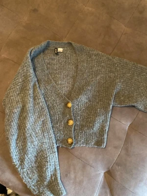 Blå cropped kofta från H&M Divided - Cropped kofta i ljusblått från H&M Divided med tre stora guldfärgade knappar framtill. Koftan har v-ringning och är stickad i ett mjukt, strukturerat material. Perfekt för att lägga till en chill vibe till din outfit.