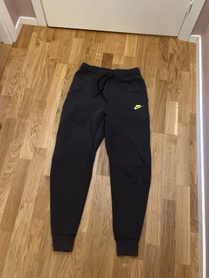 Svarta Nike mjukisbyxor med logga - Säljer ett par svarta mjukisbyxor från Nike med gul logga på vänster ben. Byxorna har dragsko i midjan, muddar vid bensluten och är tillverkade i ett mjukt och stretchigt material. Perfekta för chill eller träning.