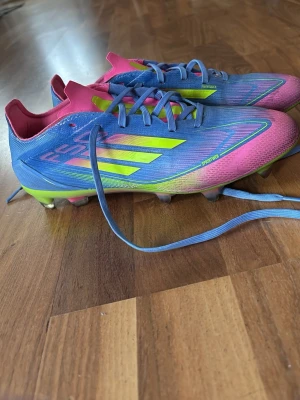 F50s - Säljer ett par färgstarka fotbollsskor från Adidas med blå ovandel, rosa detaljer och neon-gula ränder. Skorna har snörning och lätt vikt och bra grepp på planen. Har nästan aldrig andvända eftersom att de ej passade på mig så för att köpa nya vill jag sälja.