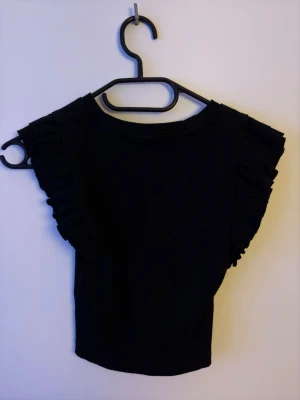 Svart volangärmad topp från Zara - Snygg svart topp från Zara med volangdetaljer längs ärmarna. Modellen är croppad och har en rund halsringning. Perfekt för dig som vill ha en enkel men ändå trendig topp till garderoben.