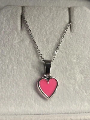 Halsband med rosa hjärthänge - Snyggt halsband i rostfritt stål med ett hjärthänge i rosa emalj som dessutom lyser i mörkret med ett lila sken. Perfekt för dig som gillar unika detaljer och vill sticka ut lite extra. Kedjan är tunn och smidig, och hjärtat har en modern, stilren design. Kedjans längd är 45 cm + 5 cm förlängningskedja. Vanligt dagsljus en kort stund räcker för att hänget ska ändra färg i mörker. 