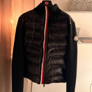 Svart cardigan från Moncler - Säljer en svart cardigan från Moncler med stickade ärmar och hög krage. Jackan har en dragkedja med röd, vit och blå rand samt Moncler-logga på ärmen. Det finns tydliga tecken på användning. Ärmarna är lite noppriga och tag saknas. Därmed reducerat pris!