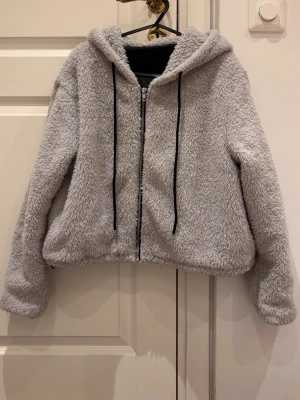 Ljusgrå teddyjacka med huva - Supermjuk ljusgrå teddyjacka med dragkedja framtill och svart snörning i huvan. Jackan har en oversized passform och fluffigt material som ger en cozy vibe. Perfekt för kyliga dagar när du vill vara både varm och stylish.
