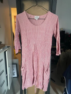 Randig röd klänning från Masai - Säljer en röd och vit randig klänning från Masai med trekvartsärm och avslappnad passform. Klänningen har rund halsringning och fickor på sidorna. Perfekt för dig som gillar en bekväm och stilren look.