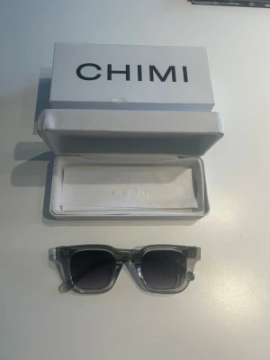 Chimi 04 Grey Solglasögon  - Snygga solglasögon från CHIMI med transparent grå båge och mörka fyrkantiga glas. Kommer i originalfodral och med putsduk. Perfekta för att lyfta din stil under soliga dagar.