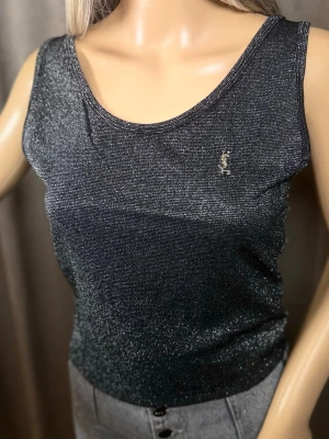 Glittrig svart linnetopp från YSL - Snygg svart linnetopp från Yves Saint Laurent med glittrigt material och ikonisk YSL-logga på bröstet. Toppen har ärmlös design och rundad halsringning, perfekt för att sticka ut med en lyxig touch.