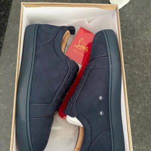 Mörkblå sneakers från Christian Louboutin - Snygga mörkblå sneakers från Christian Louboutin i mocka med klassisk röd sula och diskret broderad logga på hälen. Skorna har rund tå, snörning och beige skinnfoder för extra komfort. Perfekta för dig som vill sticka ut med exklusiv stil.