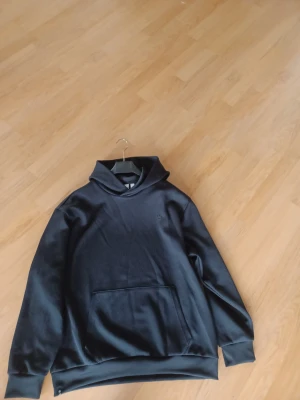 Svart hoodie Adidas  - Säljer en svart hoodie Adidas strl M med diskret ribbad struktur och stor magficka. Hoodien har huva och breda muddar vid ärmslut och nederkant. Perfekt för en chill och avslappnad stil. Bra färg, utan fel. Kan vara bara fel på min kamera. 