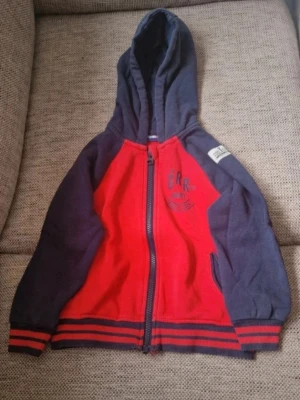 Röd och marinblå hoodie med dragkedja - Hoodie från Lupilu i rött och mörkblått. Tröjan har huva, dragkedja framtill och ribbade muddar med ränder. Tryck på bröstet med texten 'Grr not angry trouble tiger'. Utöver att färgen är något blekt vid muddarna (se bild) så hittar jag inga skavanker. Stl 98/104