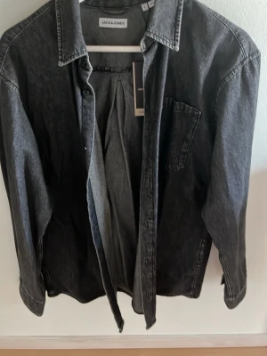 Svart jeansjacka från Jack & Jones - Snygg svart tvättad jeansjacka från Jack & Jones med klassisk krage, bröstficka och knäppning framtill. Jackan har en avslappnad passform och långa ärmar med knapp vid ärmslut. Perfekt för en cool och avslappnad stil.