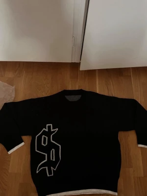 Svart stickad tröja med tryck - Cool svart stickad tröja med vitt dollartecken på framsidan och texten 'buy happiness' i gotisk stil på ryggen. Tröjan har vita detaljer vid ärmslut och nederkant. Perfekt för dig som gillar streetwear och statement-plagg.