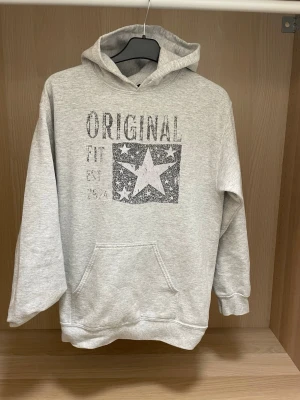 Grå hoodie - Ljusgrå hoodie med huva och magficka. Tryck på bröstet med texten 'ORIGINAL FIT' och en stor stjärna i en rektangel. Mjuk och skön, perfekt för chill dagar. Klassisk passform och ribbade muddar vid ärmar och nederkant.