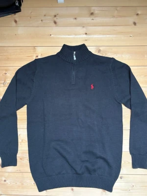 Svart stickad tröja Polo Ralph Lauren - Svart stickad tröja från Polo Ralph Lauren med halv dragkedja och ribbad hög krage. Klassisk röd logga broderad på bröstet. Perfekt för lager på lager och snygg till jeans.