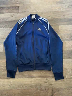 Marinblå Adidas track jacket - Snygg marinblå track jacket från Adidas med klassiska vita ränder längs ärmarna och broderad logga på bröstet. Jackan har dragkedja framtill, ribbade muddar och två fickor. Perfekt för en sportig och avslappnad stil.