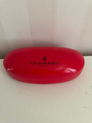 Röd solglasögonfodral Elizabeth Arden - Snyggt och stilrent rött fodral från Elizabeth Arden, perfekt för att skydda dina solglasögon. Fodralet har en hård utsida och svart insida med logga i guld. Passar de flesta solglasögonmodeller och är lätt att ta med i väskan.