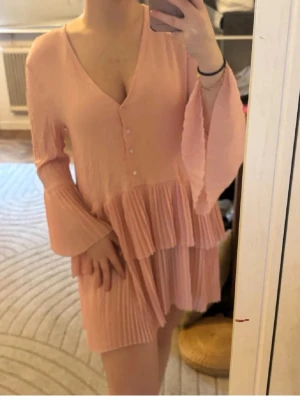Zara Ljusrosa plisserad kortklänning - Supersöt ljusrosa kortklänning med v-ringning, knappar framtill och plisserade volanger. Klänningen har långa, vida ärmar och är tillverkad i ett lätt, flowy material som ger en romantisk vibe.