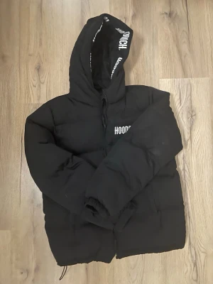Svart pufferjacka från Hoodrich - Svart pufferjacka från Hoodrich med huva och vit logga på bröstet samt tryck på insidan av huvan. Jackan har dragkedja framtill och elastiska muddar vid ärmsluten. Perfekt för kalla dagar och streetwear-stil. Xl för ungdomar 13-15y