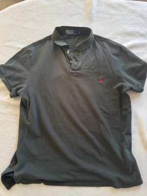 Mörkgrön Polo Ralph Lauren piké - Klassisk mörkgrå pikétröja från Polo Ralph Lauren med korta ärmar och knappar vid kragen. Tröjan har den ikoniska röda logotypen broderad på bröstet och är tillverkad i mjuk bomull. Perfekt för en avslappnad och stilren look. Står storlek L men passar mer som M