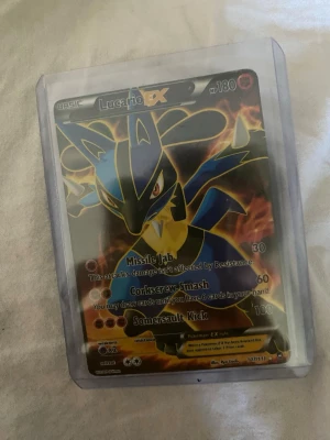 Pokémonkort Lucario EX i plastficka - Säljer ett Pokémonkort med Lucario EX, 180 HP, i en genomskinlig plastficka. Kortet har färgerna blå, gul och svart med eldig bakgrund och är från serien XY Furious Fists. Perfekt för samlare eller Pokémonfans.