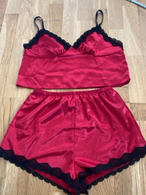 Rött satinset med spetsdetaljer - Snyggt tvådelat set i röd satin med svarta spetskanter. Setet består av ett linne med smala axelband och matchande shorts med resår i midjan. Perfekt för dig som gillar mjuka material och en lyxig känsla.