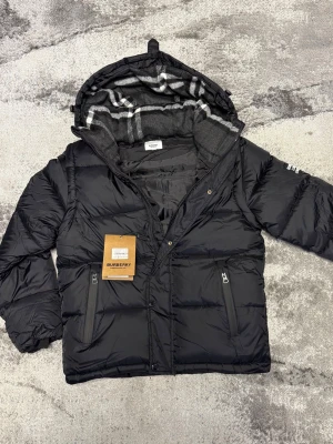 Svart pufferjacka från Burberry - Svart pufferjacka från Burberry med dragkedja, tryckknappar och två fickor framtill. Jackan har en huva fodrad med klassiskt rutmönster och är tillverkad i glansigt material. Perfekt för kalla vinterdagar och riktigt snygg streetstil.