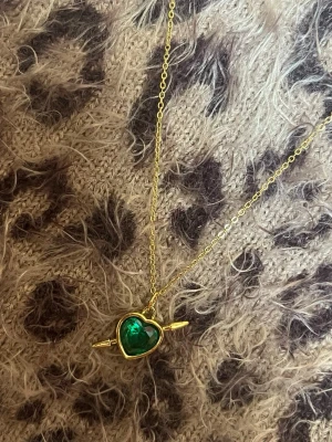 Halsband med grönt hjärthänge spjut guld - Snyggt halsband med tunn guldfärgad kedja och ett hjärtformat hänge med grön sten. Hänget har detaljer i guld runt stenen och en pil som går genom hjärtat. Perfekt accessoar för att lyfta din outfit. Rostfri 
