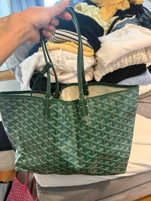 Grön Goyard väska - Snygg grön väska från Goyard med klassiskt mönster i vitt och mörkgrönt. Väskan har två långa handtag och är tillverkad i canvas med läderdetaljer. Perfekt för dig som vill ha en rymlig och trendig väska med ikonisk design.