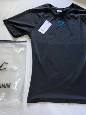 Gymshark onyx V5 compression shirt  - Svart compression shirt från Gymshark med blå logga på bröstet. Tillverkad i ett lätt och syntetiskt material som andas, perfekt för intensiva träningspass. Modellen tillverkas inte längre så den är sällsynt. Snygg sportig design med diskret mönster på övre delen. Strl S men sitter som M. Hör av er vid intresse eller frågor!