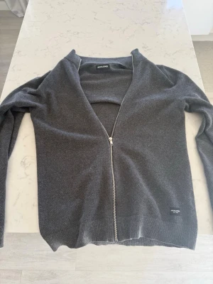 Mörkgrå ziptröja från Jack & Jones - Snygg mörkgrå stickad ziptröja från Jack & Jones med hel dragkedja framtill och liten logotyp nertill. Tröjan har en enkel, rak passform och är tillverkad i ett mjukt material. Perfekt att slänga på sig över en t-shirt.