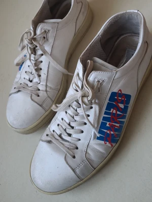 Vita Kenzo Paris sneakers - Säljer ett par vita Kenzo sneakers i läder med rött Paris-tryck på sidan. Klassisk låg modell med vita snören och gummisula. Perfekta för dig som gillar streetstyle och vill ha något unikt från ett känt märke. Använda, men inga defekter som påverkar funktion. Lite slitna, men inget slitage. Några små smutsfläckar som syns på nära håll.
