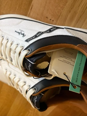 Vita sneakers från Pepe Jeans - Stilrena vita sneakers från Pepe Jeans med detaljer i mörkblått och brunt. Skorna har textilovandel, vita snören och diskret logga på sidan. Snygg kontrast mellan färgerna och klassisk låg modell som passar till många outfits. Strl 41/42