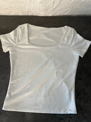 Vit topp med fyrkantig ringning - Snygg vit topp med korta ärmar och fyrkantig ringning. Tunn och stretchig, perfekt att styla med jeans eller kjol. Enkel och stilren design som passar till många looks.