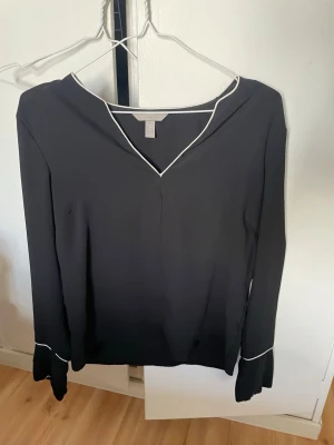 Svart blus med vita detaljer H&M - Stilren svart blus från H&M med vita kantdetaljer vid halsringning och ärmslut. Blusen har v-ringning och långa ärmar, perfekt för en clean och modern look. Materialet känns lätt och smidigt, vilket gör den enkel att bära till många outfits.