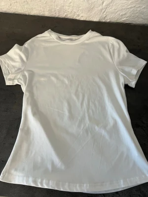 Vit basic t-shirt i bomull - En klassisk vit t-shirt i mjuk bomull med rund hals och korta ärmar. Perfekt att matcha med jeans eller shorts för en clean och enkel stil. T-shirten har en rak passform och är helt utan tryck eller mönster.