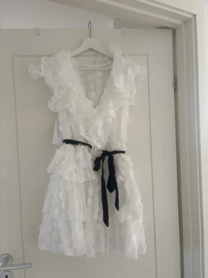 Vit volangklänning med knytband - Supersöt vit kortklänning med volanger och v-ringning. Klänningen har ett svart knytband i midjan och är ärmlös. Tyg med diskret struktur och flera lager ger en romantisk look. Perfekt för sommarens alla tillfällen och även en perfekt studentklänning som matchar mössan fint med de svarta bandet. 
