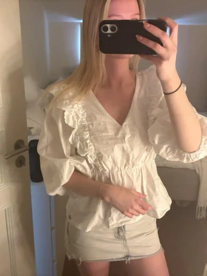 Vit blus med volanger och v-ringning - Snygg vit blus med v-ringning och volangdetaljer längs axlar och bröst. Blusen har trekvartsärm och är tillverkad i ett lätt, luftigt tyg som ger en romantisk känsla. Perfekt att matcha med jeans eller kjol för en trendig look.