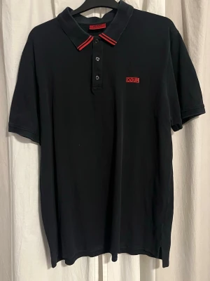 Svart pikétröja från Hugo Boss - Svart pikétröja från Hugo Boss med röd logga broderad på bröstet och röda ränder på kragen. Klassisk polokrage med tre knappar framtill. Perfekt för en stilren och sportig look.