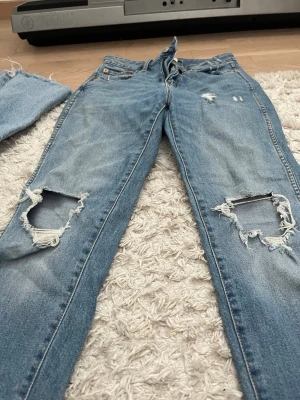 Ljusblå ripped jeans med hål - Säljer ett par ljusblå jeans med stora slitna hål på båda knäna och distressed detaljer framtill. Jeansen har klassisk femficksdesign och raka ben. Perfekta för en avslappnad och trendig streetstil.