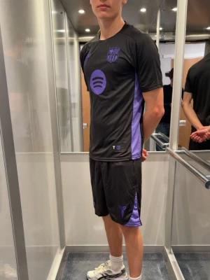 FC Barcelona träningsset - Säljer ett svart och lila träningsset från FC Barcelona. Setet består av en t-shirt och shorts i lätt och snabbtorkande material, perfekt för träning eller match. Både tröjan och shortsen har klubbens logga och lila detaljer på sidorna.
