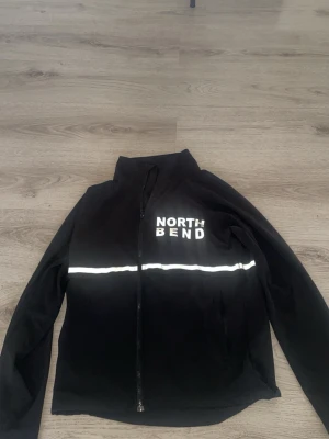 Svart vindjacka från North Bend - Svart vindjacka med hel dragkedja och vit reflexdetalj runtom bröstet. Jackan har hög krage och logga på bröstet samt reflexmärke bak. Perfekt för blåsiga dagar och kvällspromenader.