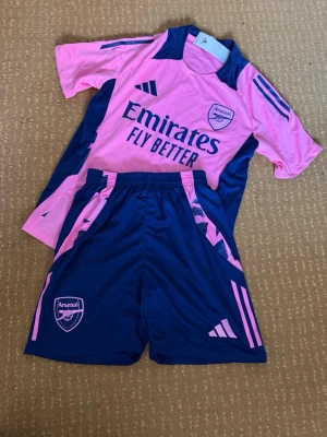 Arsenal träningsset  - Säljer ett Arsenal träningsset från Adidas med rosa t-shirt och marinblå shorts. Tröjan har mörkblå detaljer och texten 'Fly Better', medan shortsen har rosa ränder och Arsenal-logga. Perfekt för fotbollsträning eller gymmet.