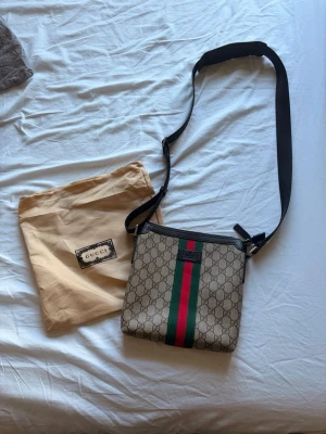 Gucci axelväska  - Snygg axelväska från Gucci i beige canvas med klassiskt GG-monogram och gröna samt röda ränder framtill. Väskan har svart justerbar axelrem och svarta detaljer. Perfekt för dig som gillar lyxiga accessoarer med ikonisk design.