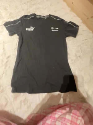 Svart Puma BMW Motorsport t-shirt - Säljer en svart Puma t-shirt i samarbete med BMW Motorsport. T-shirten har vita loggor på bröstet och tunna vita detaljer längs axlarna. Klassisk passform och rund hals. Perfekt för dig som gillar motorsport och streetwear.