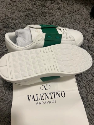 Valentino Garavani vita sneakers - Snygga vita sneakers från Valentino Garavani med en grön bred rem över mitten och matchande gröna nitar. Skorna är i läder och har klassisk låg siluett med vita snören och diskret logga på plösen. Perfekta för en clean och trendig look. Storlek 44 men passar oxå för 43