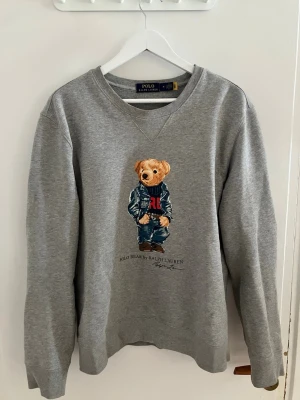 Grå sweatshirt Polo Bear Ralph Lauren - Grå sweatshirt från Ralph Lauren med ikoniska Polo Bear-trycket framtill. Tröjan har rund halsringning och är tillverkad i mjukt bomullsmaterial. Perfekt för en avslappnad och cool stil.