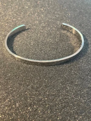 Silvrigt minimalistiskt armband - Ett stilrent och snygg herr armband i silverfärgad metall. Armbandet är stelt och öppet, med en slät och blank yta som ger en modern känsla. Perfekt för dig som gillar enkel och tidlös design. priset är lite diskuterbart