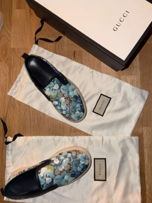 Gucci slip-on sneakers med blommor - Slip-on sneakers från Gucci med blått och grönt blommönster på canvas, vita gummisulor och svarta läderdetaljer. Snygg och unik design som sticker ut, perfekt för dig som gillar statement-plagg och exklusiva märken.