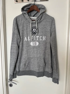 Grå hoodie från Abercrombie & Fitch - Snygg grå hoodie från Abercrombie & Fitch med broderad logga och text framtill. Klassisk känguruficka, dragsko i huvan och ribbade muddar. Perfekt för en avslappnad och sportig stil. Mjuk bomullsblandning för extra komfort.