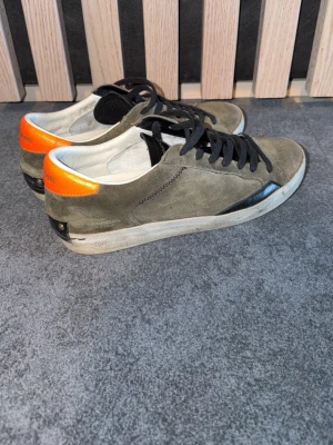 Gröna sneakers från Crime London - Stilrena sneakers i mocka från Crime London med olivgröna paneler, svarta detaljer och en orange hälkappa. Skorna har svarta skosnören och vit sula. Perfekta för dig som gillar en urban och trendig look. Skick 8/10 hör av dig vid frågor. Box ingår🤗 storlek 41 men passar även 42
