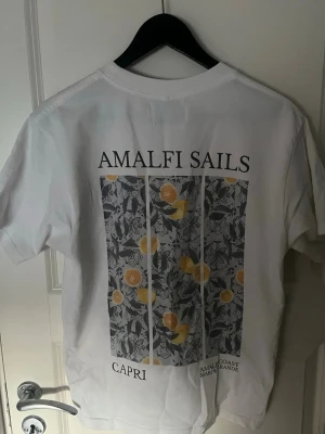 Notre Succes T-shirt - Amalfi Sails tryck - Vit t-shirt från Notre Succes med text på bröstet och stort Amalfi Sails-tryck på ryggen med citronmotiv. Klassisk passform och rund hals. Perfekt för dig som gillar stilrena plagg med snygga detaljer.